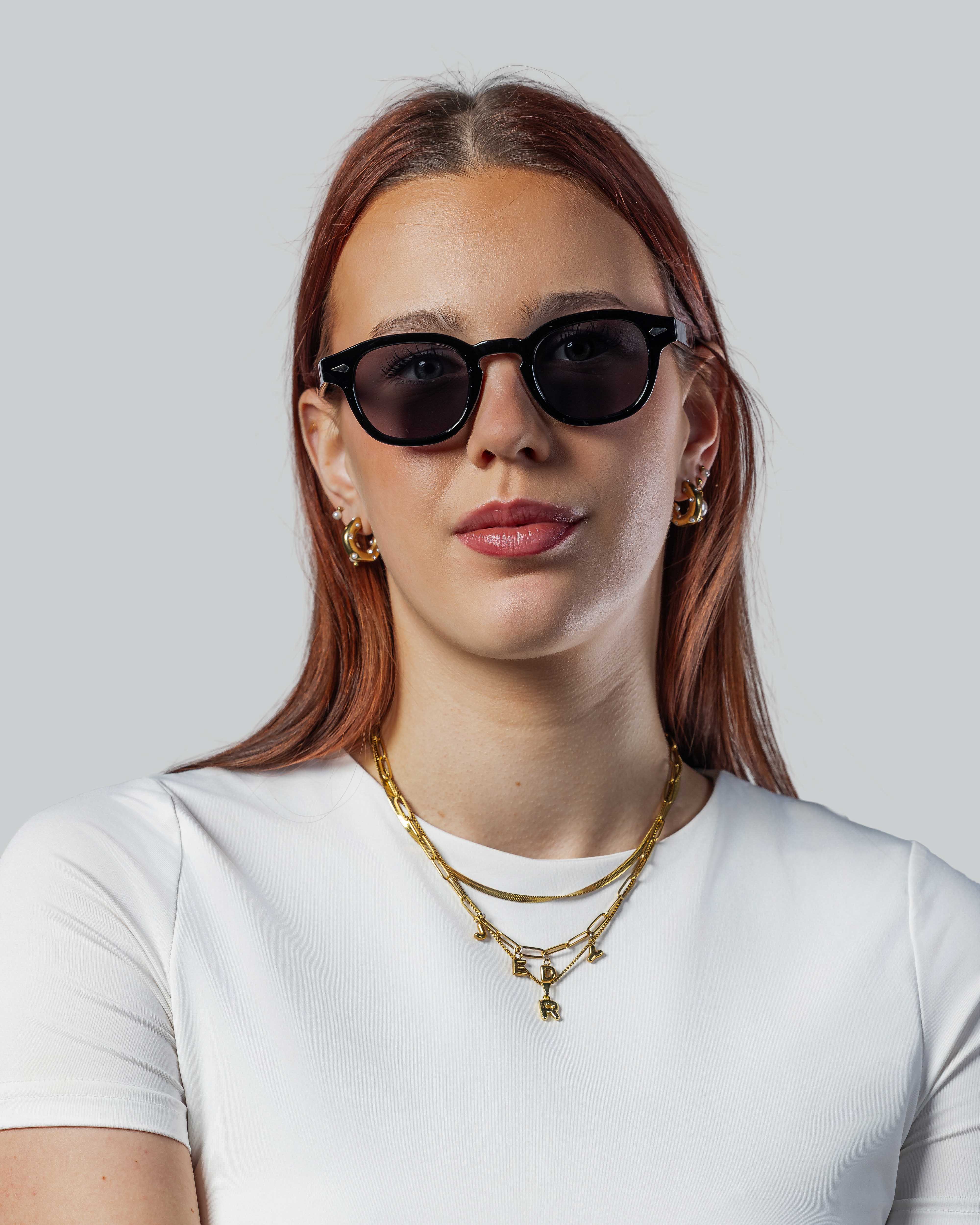 Vrouw met stijlvolle zwarte zonnebril, gouden sieraden en witte top, perfect voor zomerse uitjes.