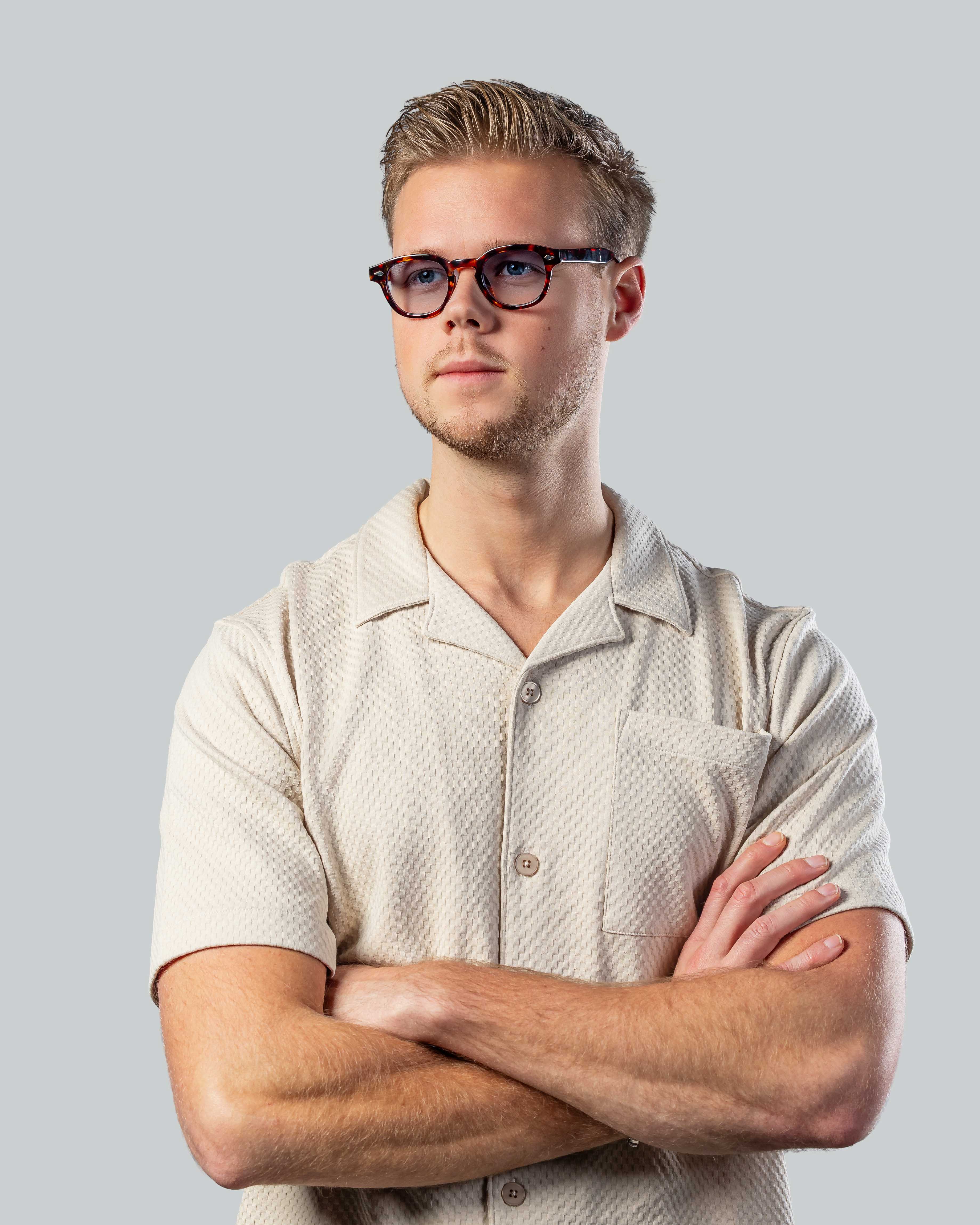 Man met bril en beige overhemd, armen over elkaar, stijlvolle zonnebril