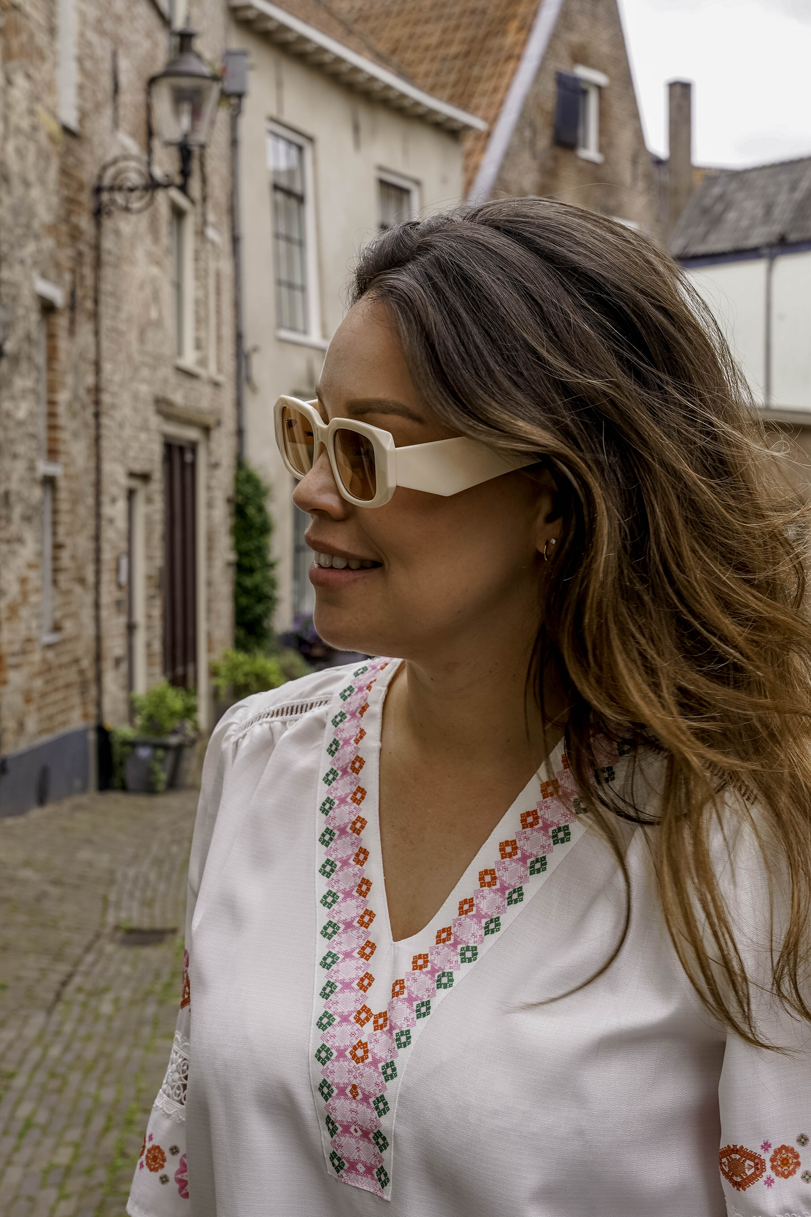 Vrouw met stijlvolle witte zonnebril en geborduurde blouse in oude straat
