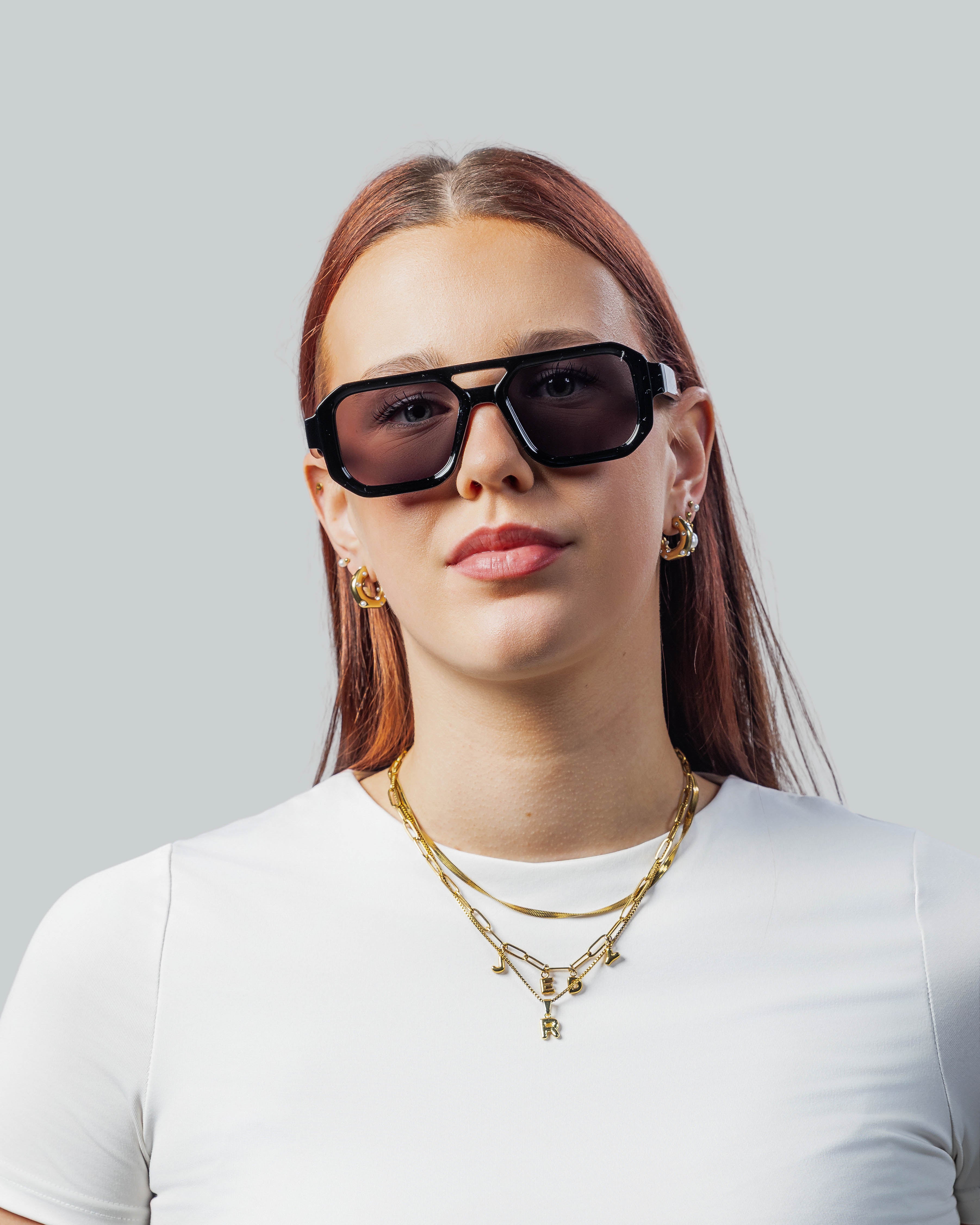 Stijlvolle zwarte zonnebril met grote montuur, gedragen door vrouw met gouden sieraden en wit shirt