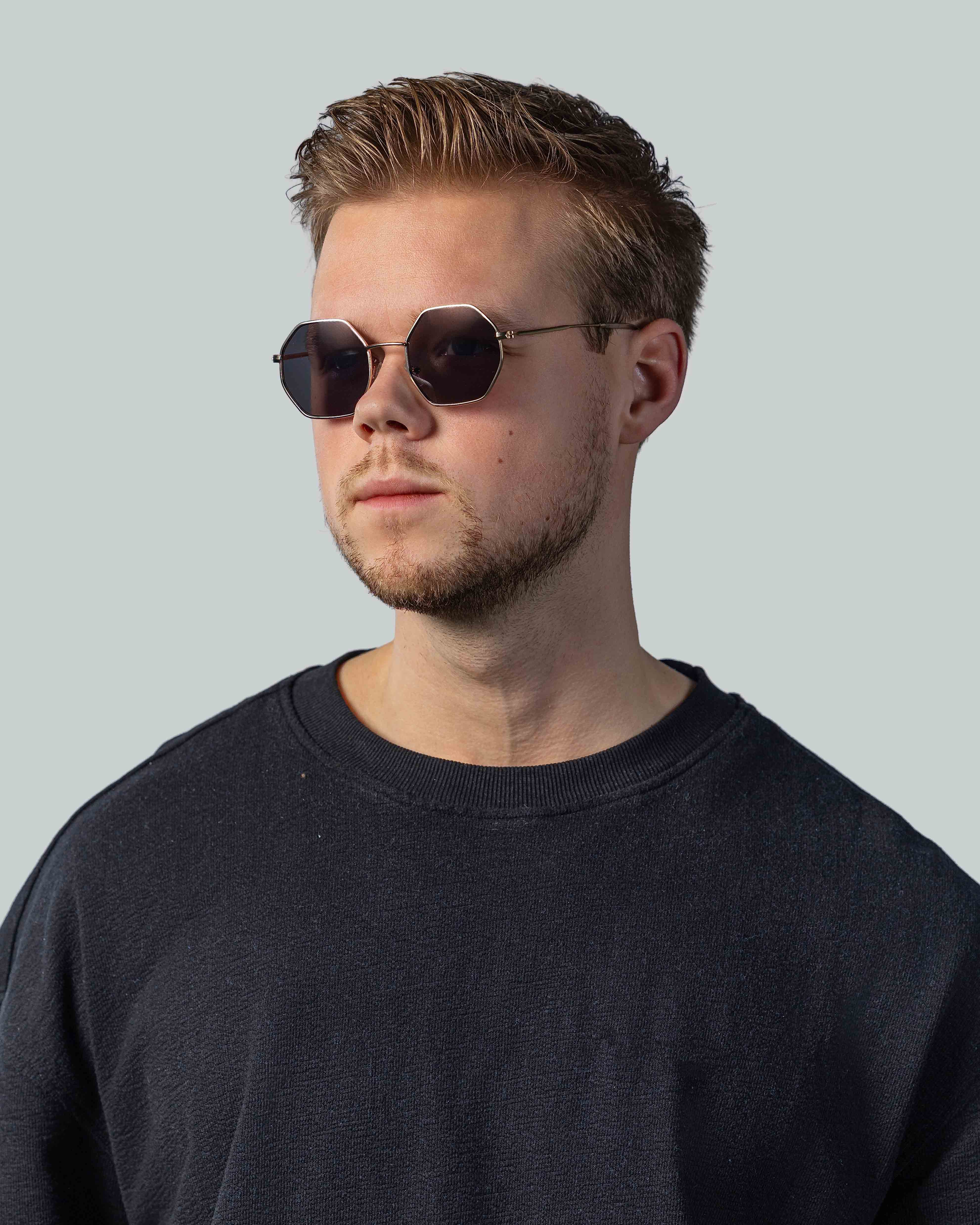 Stijlvolle zonnebril met octagonale glazen en gouden montuur, gedragen door man in donkere sweater.