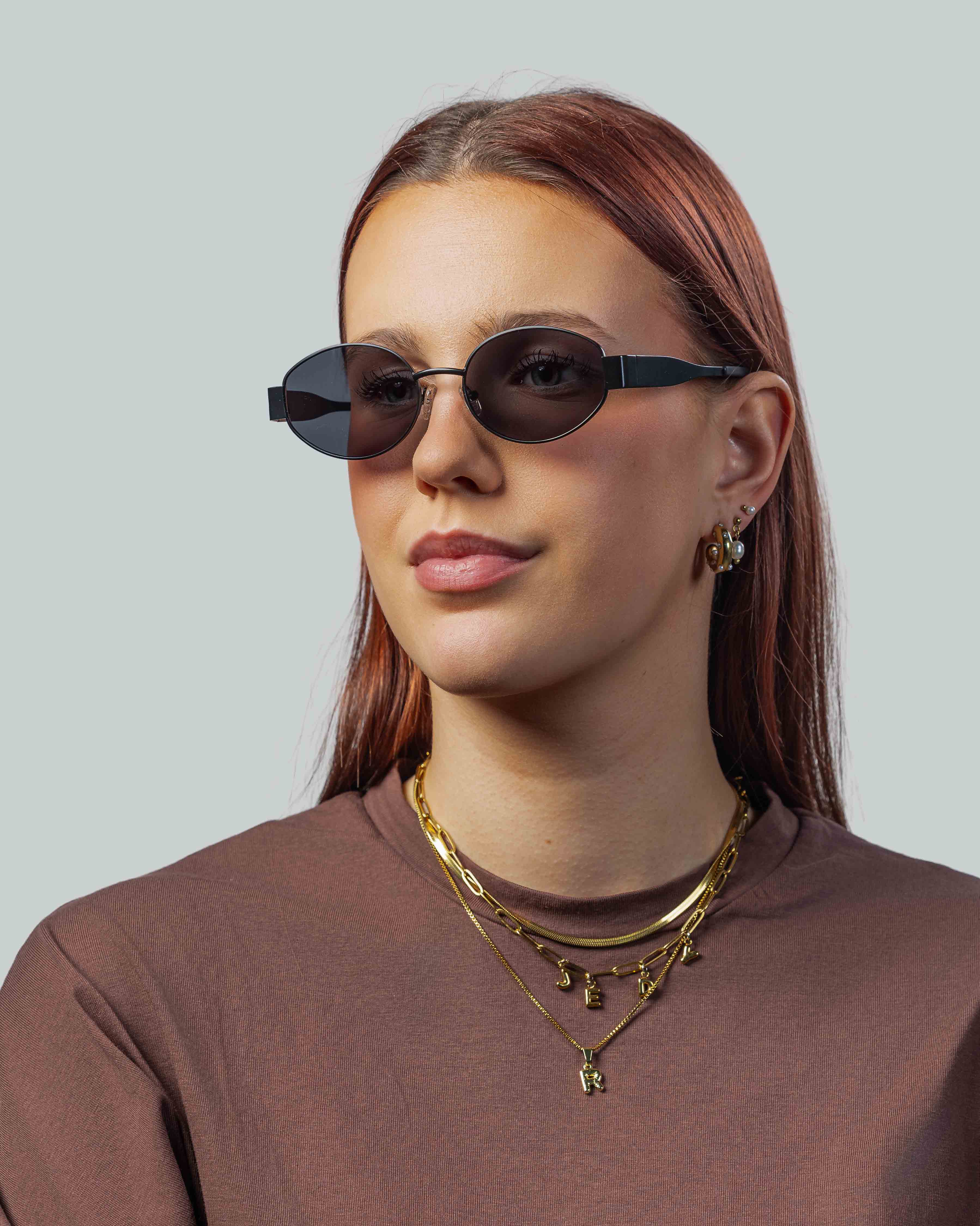 Stijlvolle ovale zonnebril met donkere glazen en zwarte montuur, gedragen door vrouw met gouden sieraden