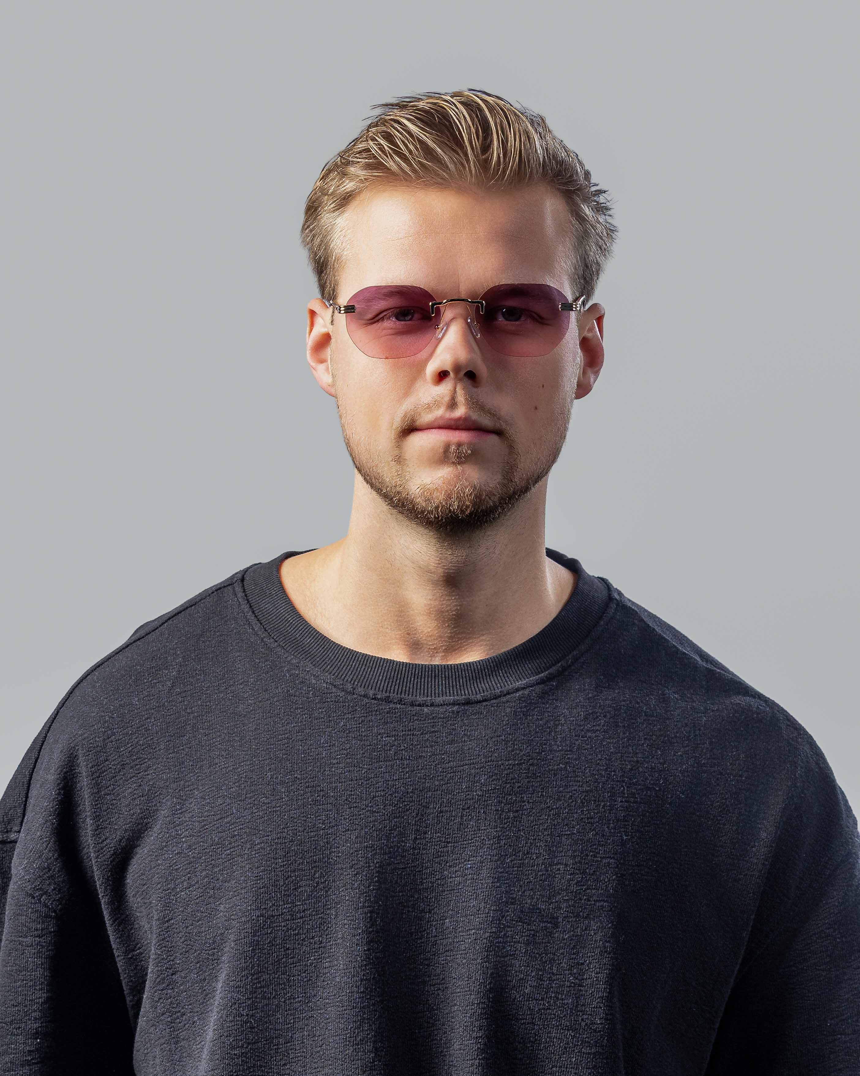 Stijlvolle zonnebril met roze glazen en goudkleurige montuur, gedragen door man in donkere sweater.