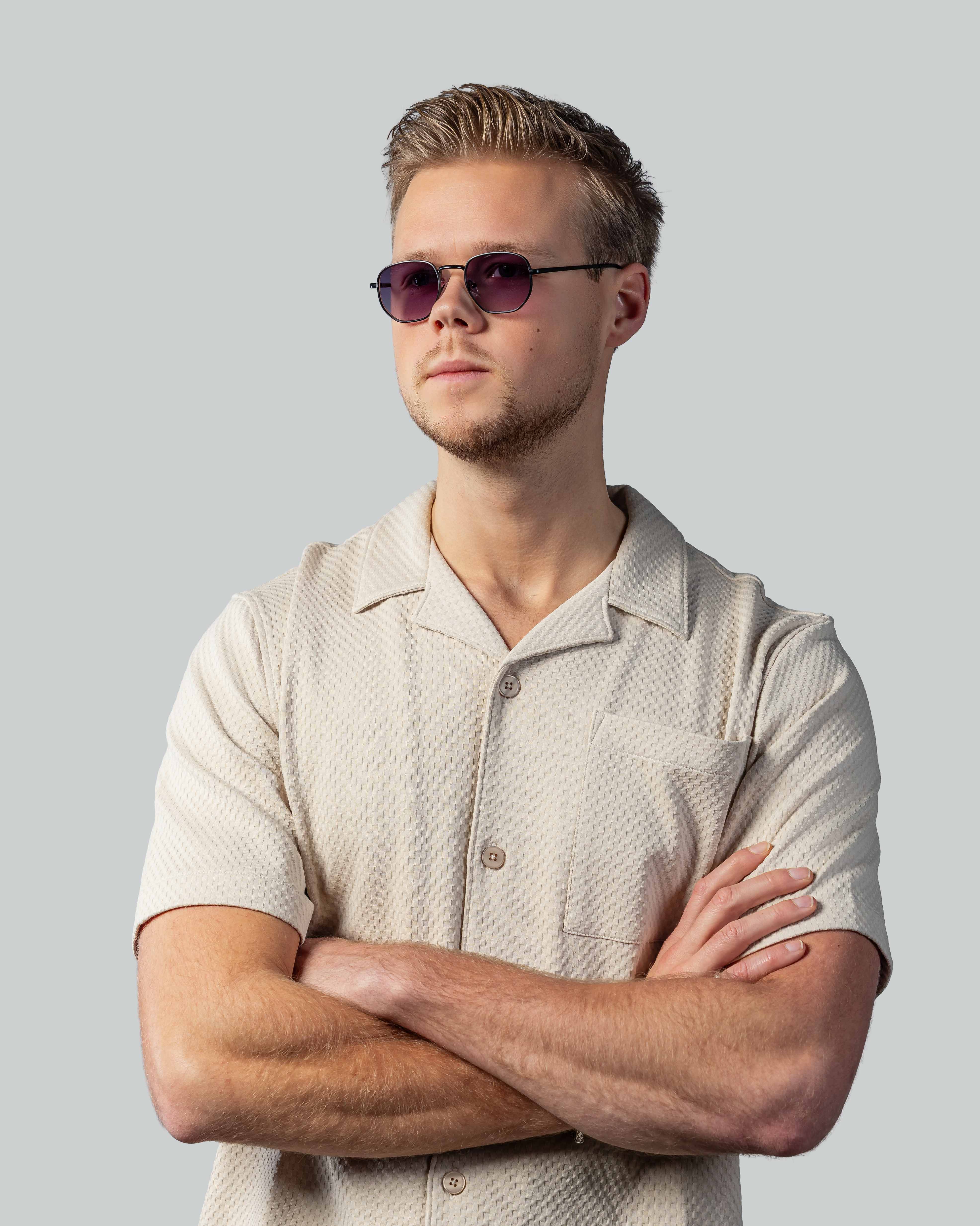 Stijlvolle zonnebril met paarse glazen en zwarte montuur, gedragen door man in beige shirt met armen over elkaar.