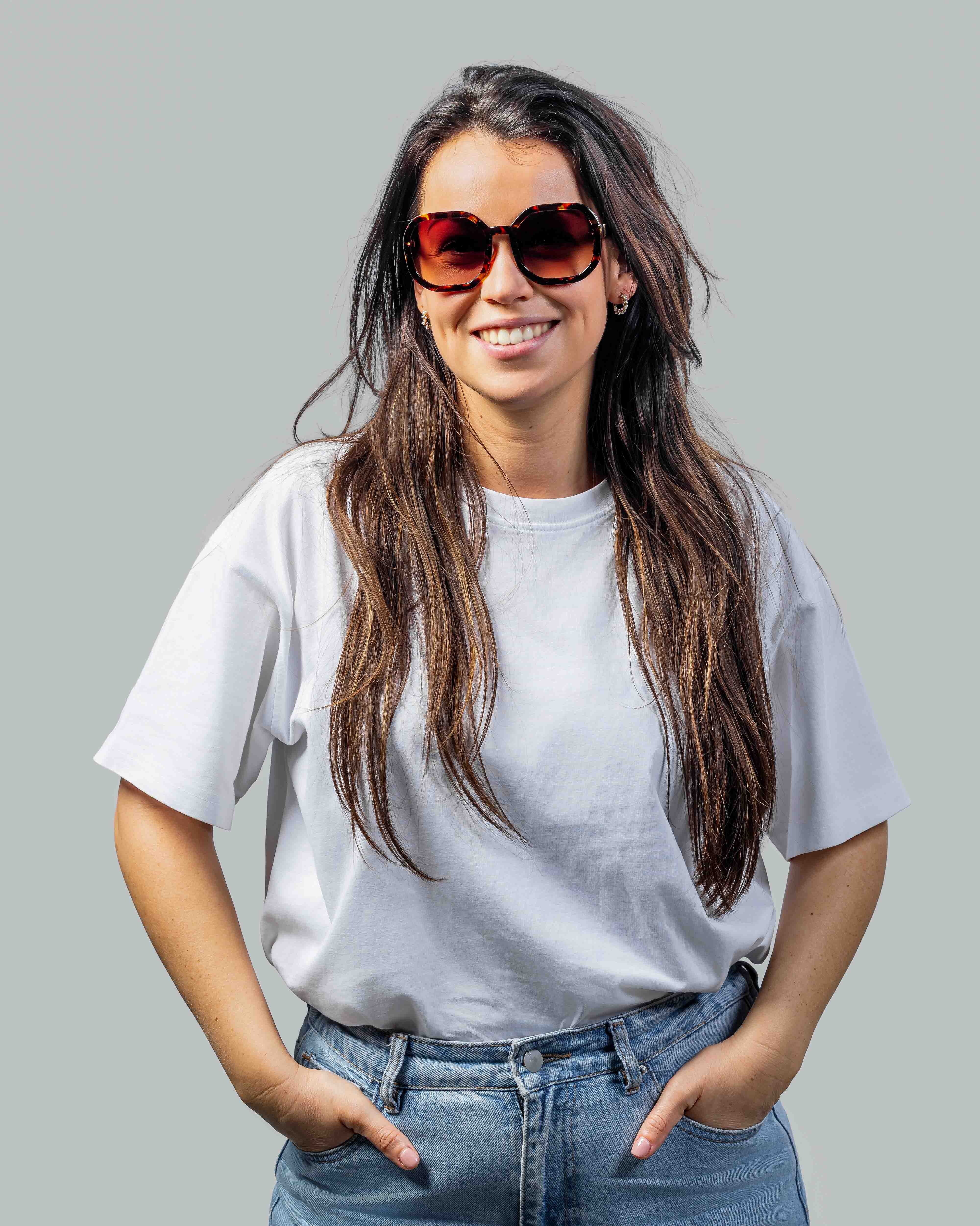 Vrouw met stijlvolle zonnebril, wit T-shirt en jeans, glimlachend met handen in zakken
