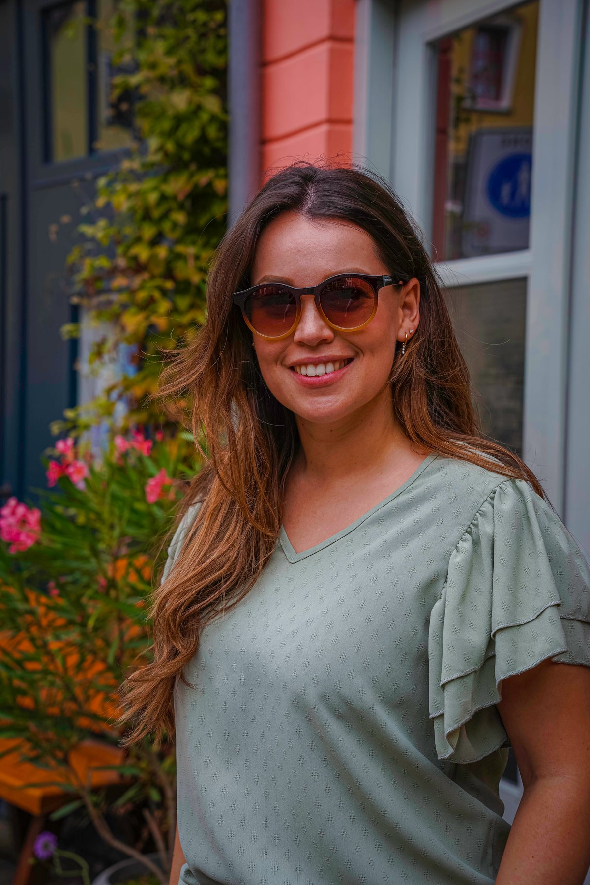 Vrouw met stijlvolle zonnebril en groene blouse, glimlachend buiten bij bloemen