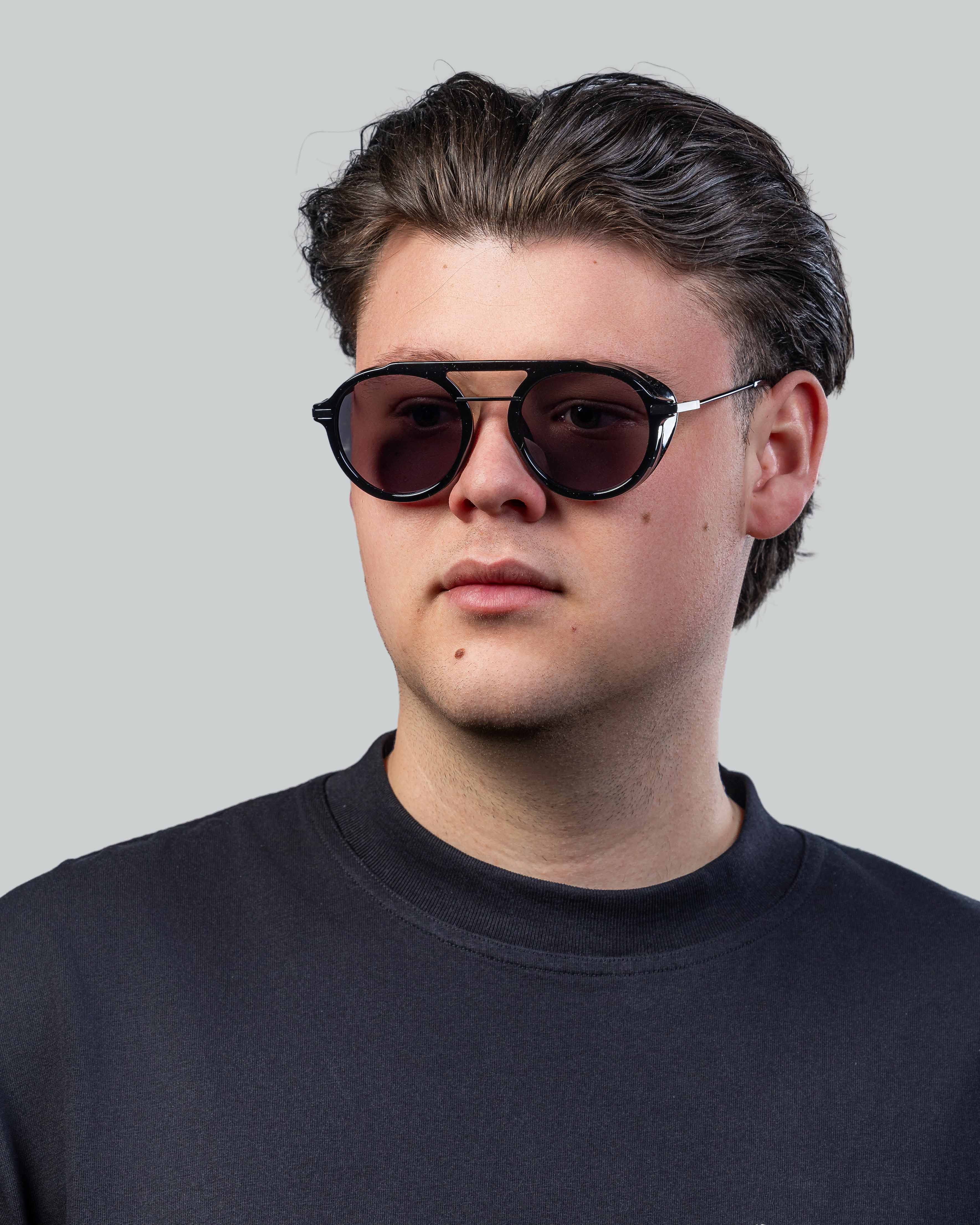 Stijlvolle zonnebril met zwarte montuur en donkere glazen, gedragen door man in donkergrijs shirt