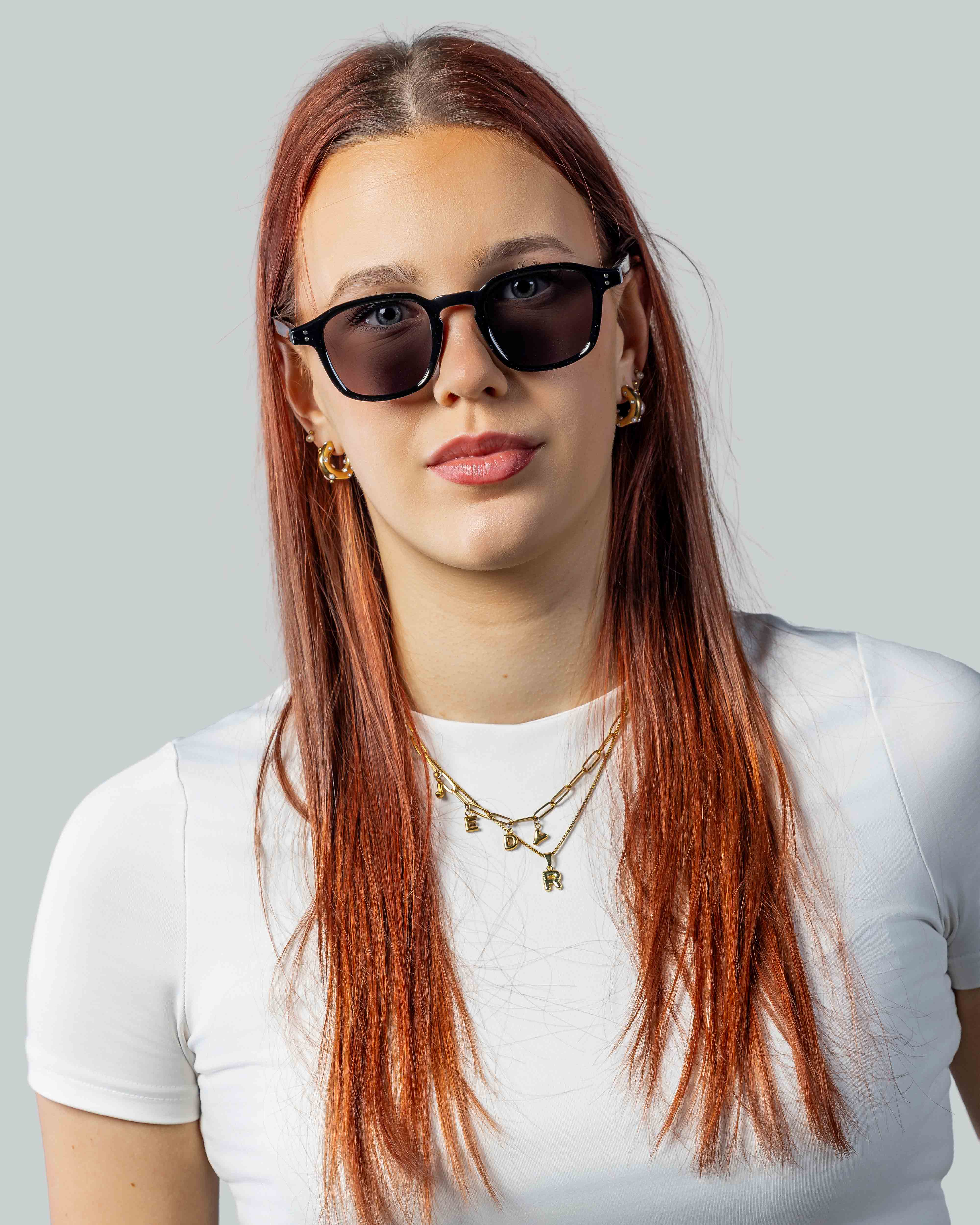 Vrouw met stijlvolle zwarte zonnebril, gouden sieraden en witte top tegen lichte achtergrond